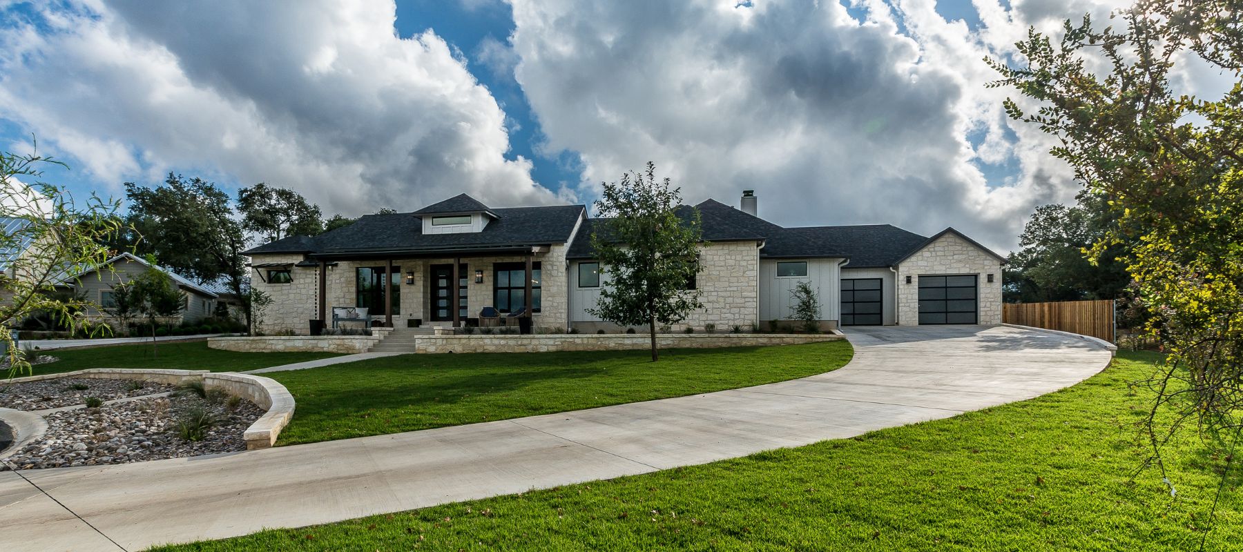 Spacious suburban home with a well-maintained green lawn and a curved driveway leading to a two-car garage. The house features a stone exterior, a covered porch with seating, and large windows. The sky above is partly cloudy, providing a picturesque backdrop to the serene residential scene.