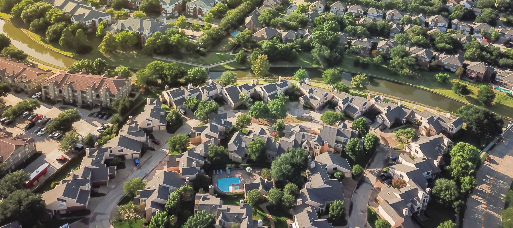 Aerial view of a residential neighborhood featuring a collection of houses with gray roofs, surrounded by green trees and landscaping. The image also includes a swimming pool and a winding street layout. In the background, there are additional homes and a small body of water, all set within a lush, green environment