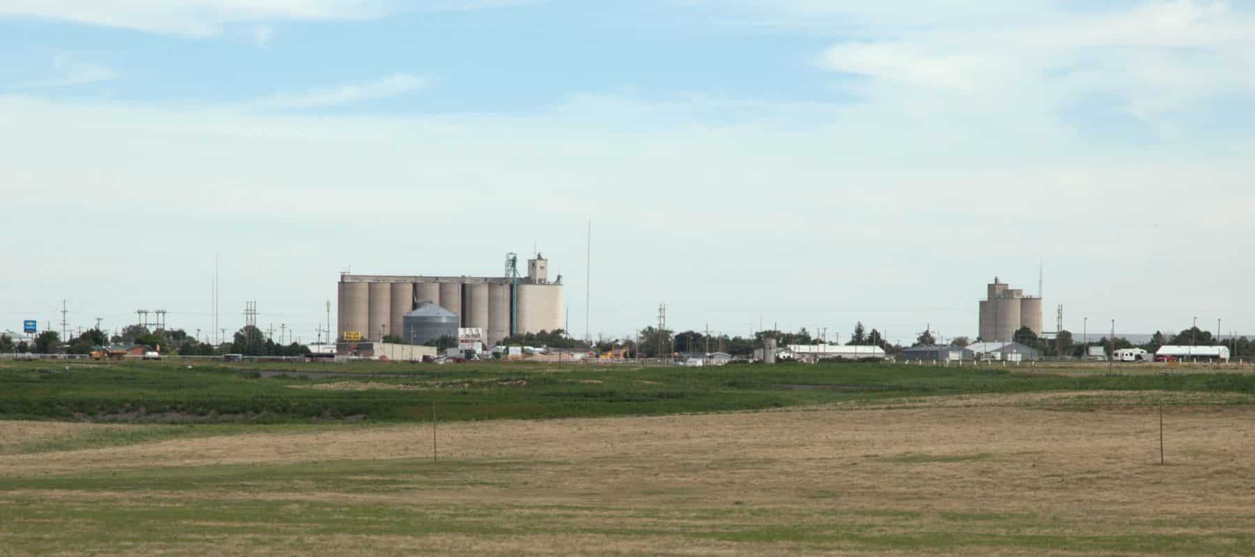 Wide view of a rural landscape with large grain silos and agricultural buildings in the distance. The foreground features open fields with a mix of grass and dirt, while the sky above is partly cloudy, highlighting the expansive countryside