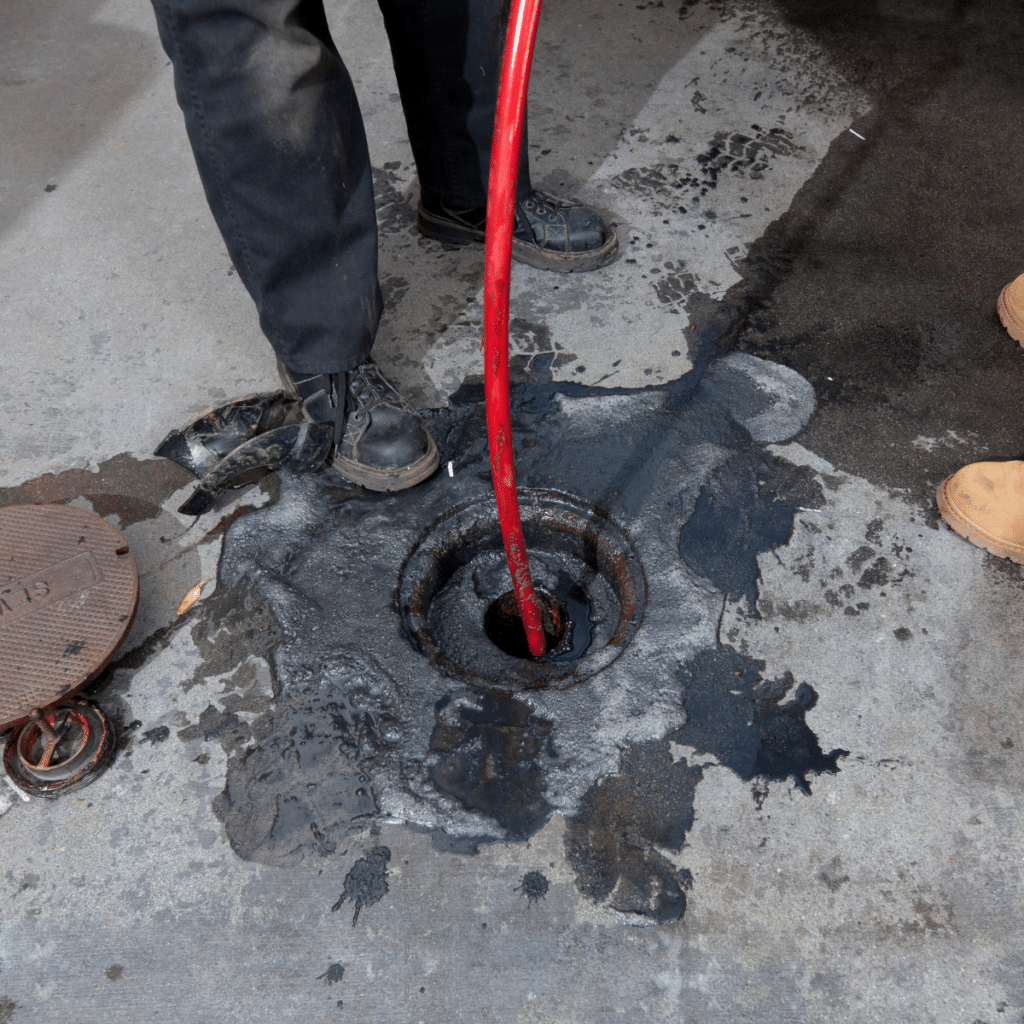 A worker performing hydro jetting, using a red hose to clear a clogged drain surrounded by water and debris