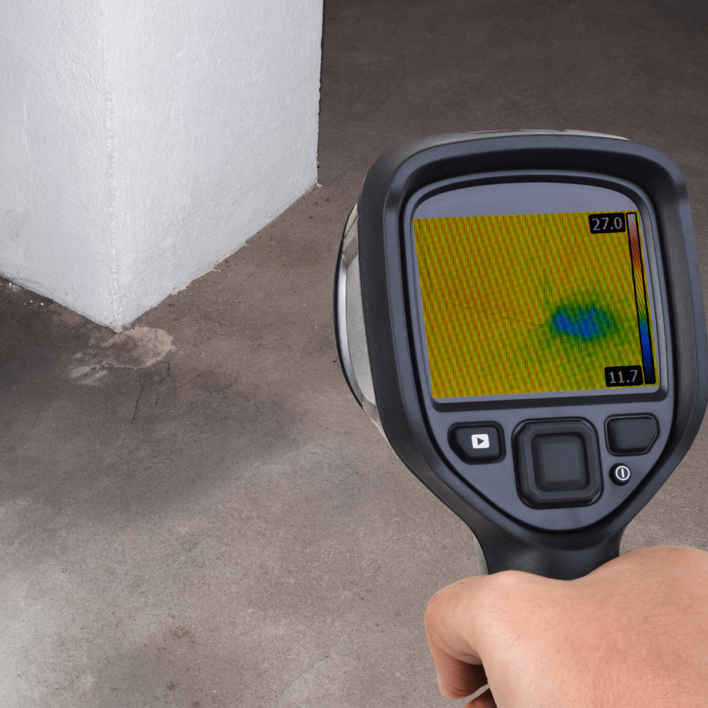 A handheld thermal imaging camera detecting a water leak on a concrete floor, showing a blue and green heat signature