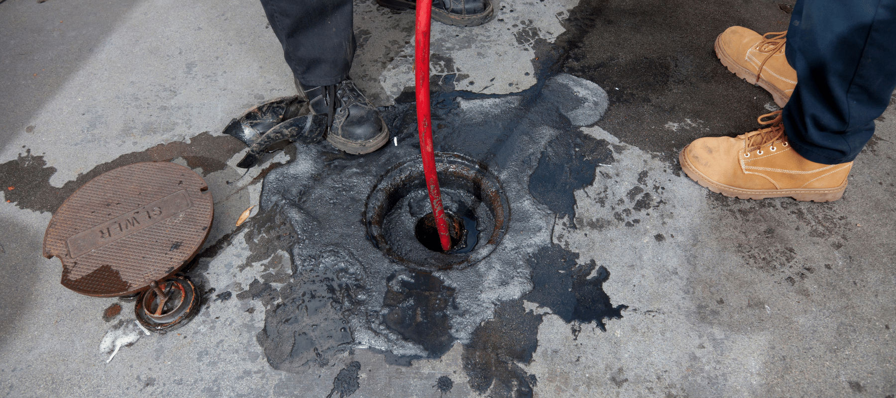 A worker performing hydro jetting, using a red hose to clear a clogged drain surrounded by water and debris