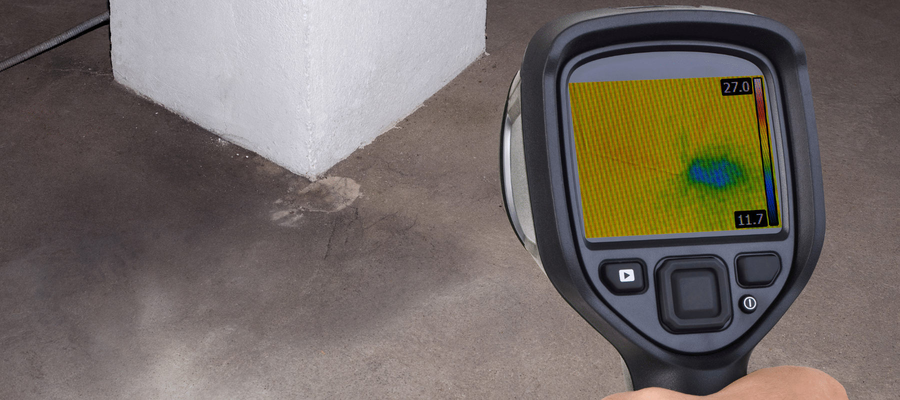 A handheld thermal imaging camera detecting a water leak on a concrete floor, showing a blue and green heat signature