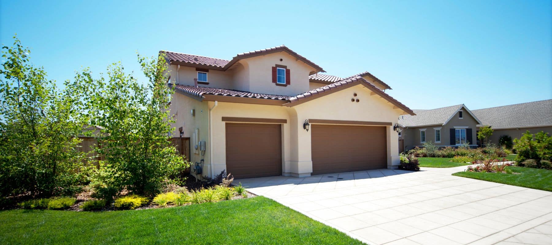 A large, two-story stucco house with a reddish-brown tile roof and two brown garage doors sits under a clear blue sky, surrounded by green lawns and trees