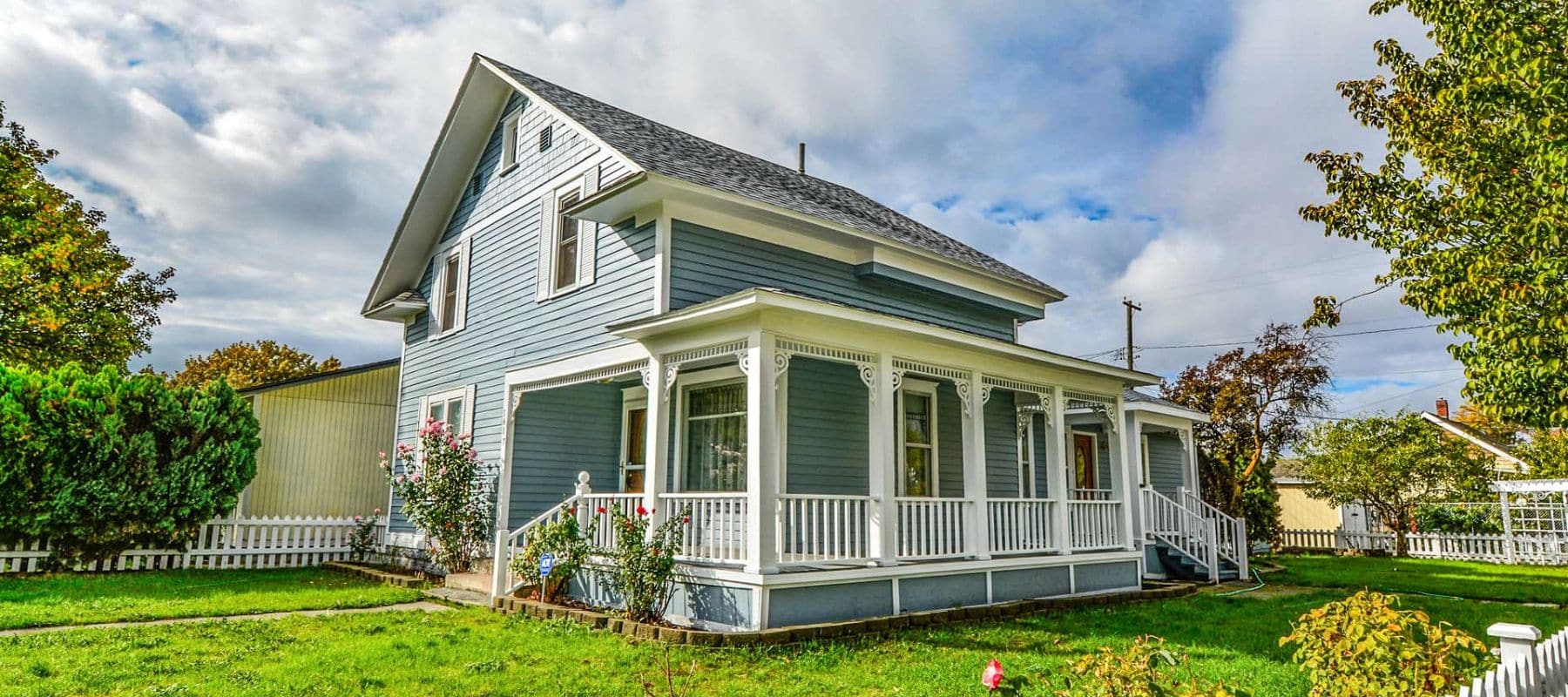A charming light blue two-story house with white trim and a wrap-around porch stands proudly on a green lawn under a partly cloudy sky