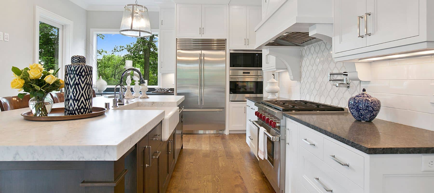 A spacious and bright modern kitchen boasts white cabinetry, a large stainless steel refrigerator, a range, and marble countertops with a sink, all beneath a large window