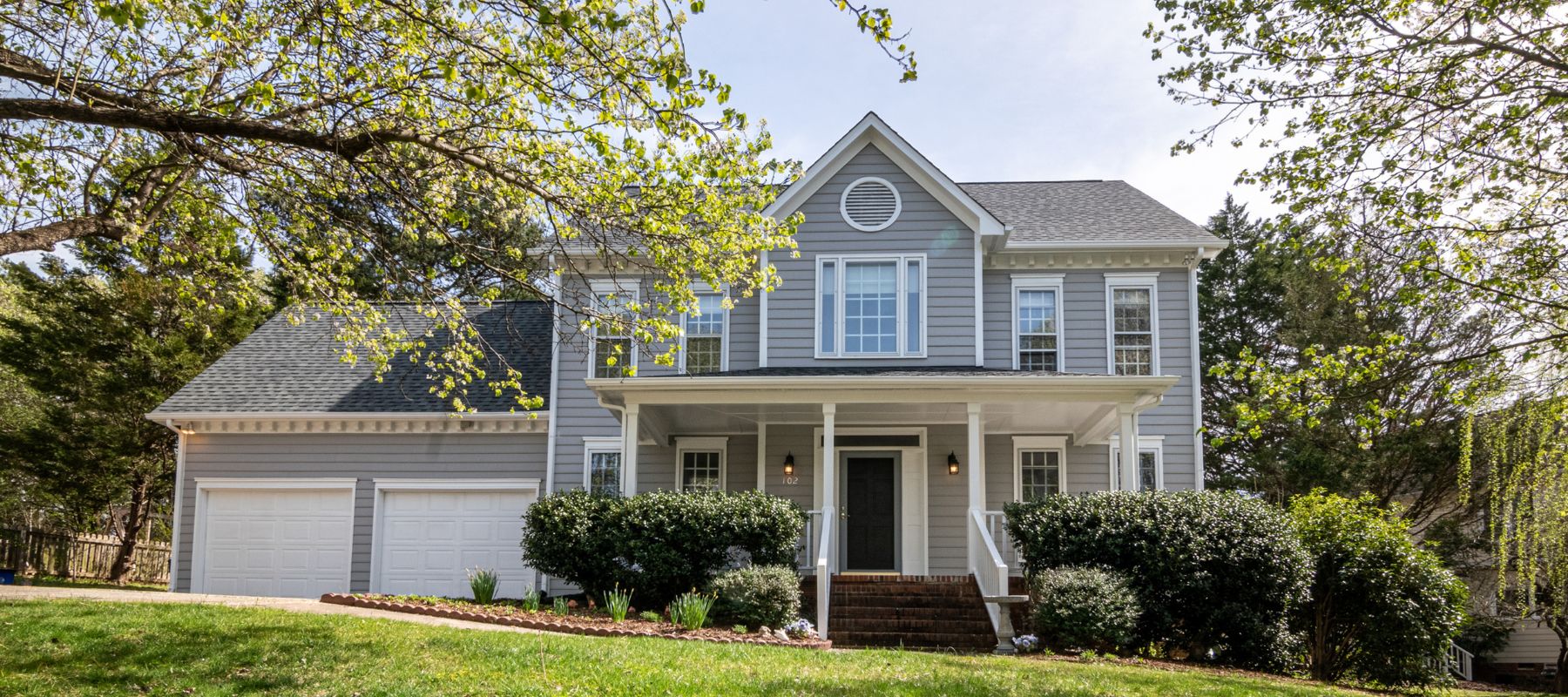 A stately gray two-story house with white trim features a welcoming front porch, a two-car garage, and is surrounded by lush green trees and shrubs under a bright sky