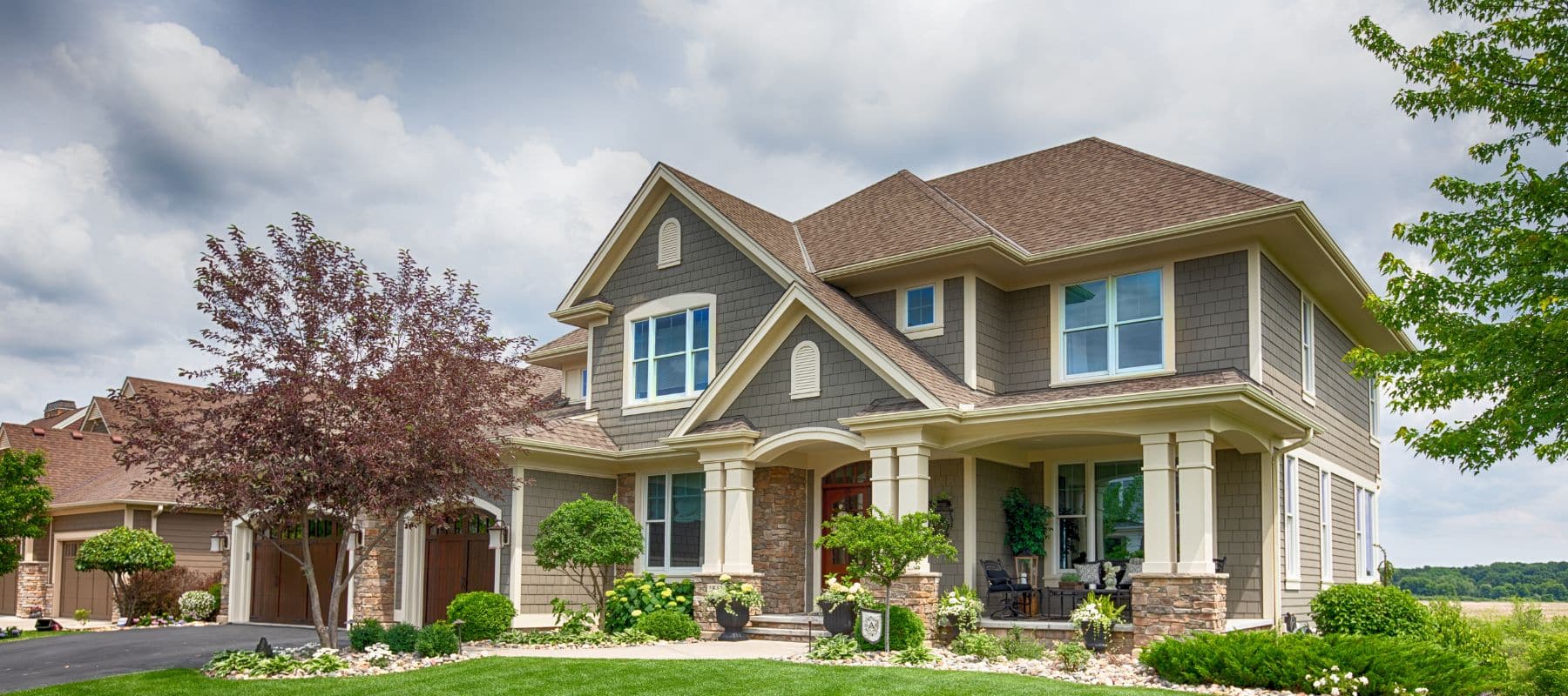 A grand, modern two-story house with a mix of brown siding and stone accents features a large front porch and well-maintained landscaping under a cloudy sky