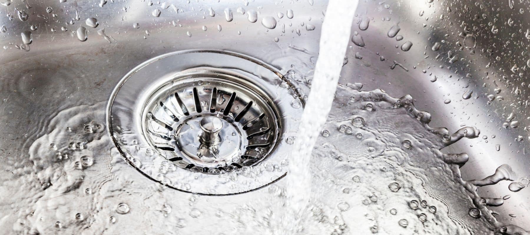 Water running into a stainless steel sink drain with droplets splashing