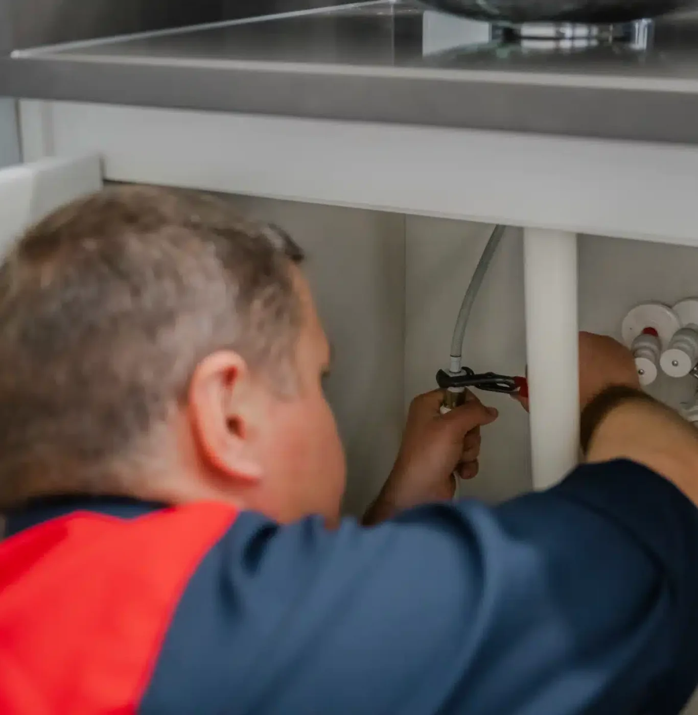 A plumber is seen from behind, using a tool to work on the pipes and connections under a kitchen sink