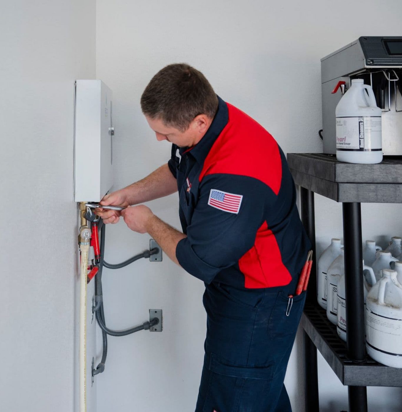 A plumber adjusts a connection on a wall-mounted tankless water heater next to a utility shelf